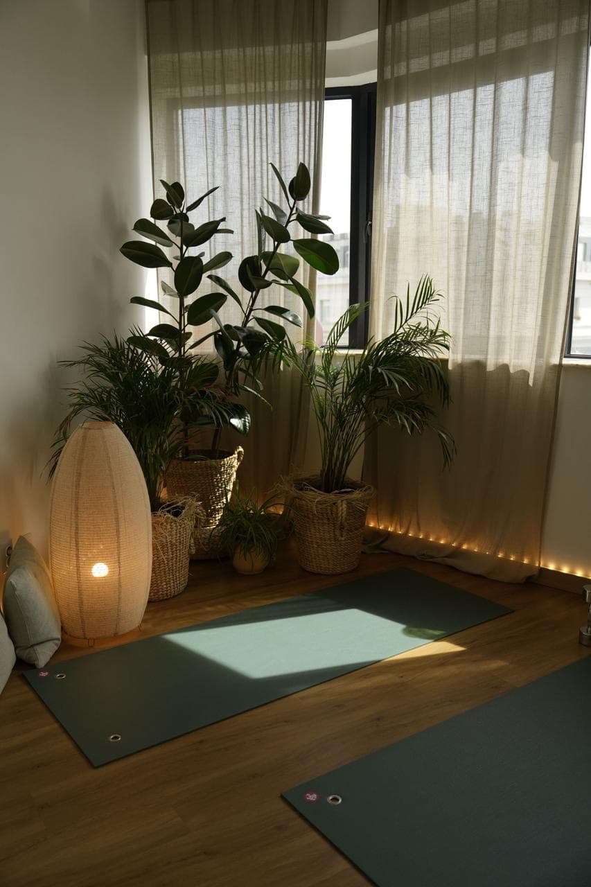 A cozy, dimly lit corner of the studio featuring a tall oval floor lamp, large potted plants in woven baskets, and a yoga mat on the wooden floor.